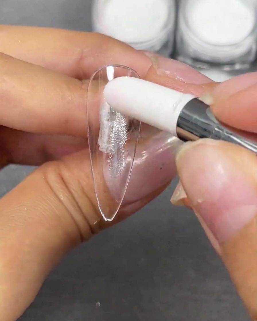 Une main utilisant un applicateur mousse blanc pour tamponner de la poudre argentée Crystal Plating sur une capsule dongle stiletto claire.