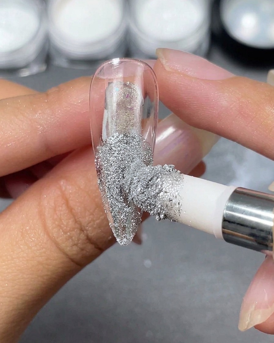Un applicateur en mousse déposant de la poudre chrome argentée sur une capsule dongle stiletto transparente tenue entre les doigts.