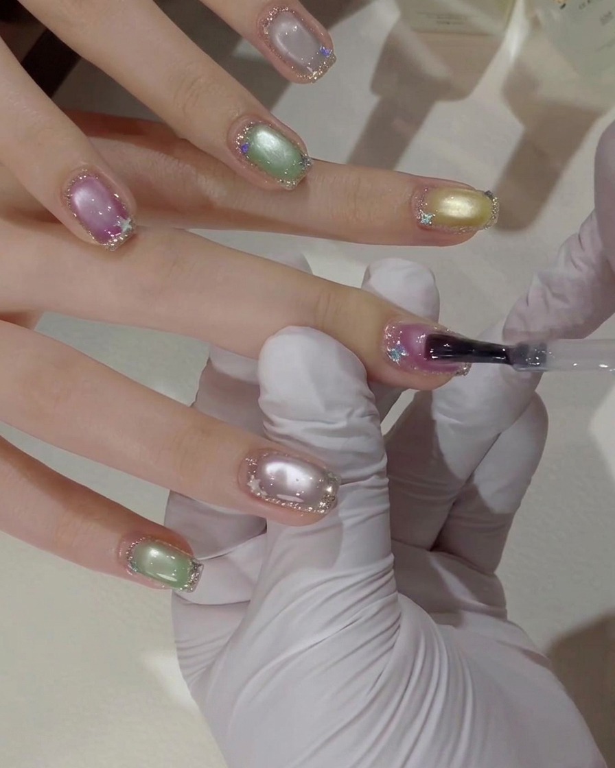Gros plan dune main gantée utilisant un pinceau à gel pour appliquer une couche de finition transparente sur un ongle rose et violet effet œil de chat avec des bordures à paillettes argentées.