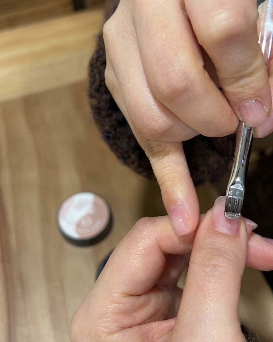 Gros plan dun pinceau à gel appliquant un vernis nude doré scintillant sur un ongle naturel.