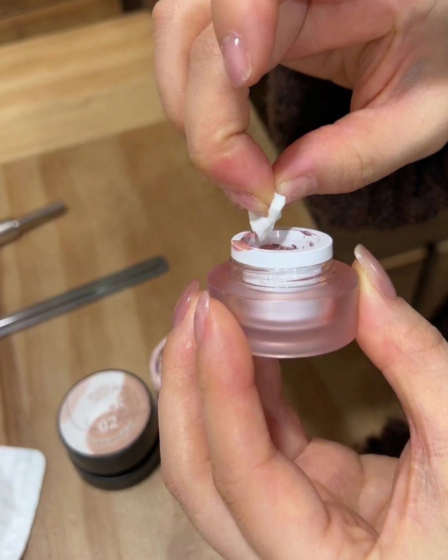 Gros plan de mains utilisant un morceau déchiqueté déponge de maquillage blanche pour prélever du vernis gel rose dans un petit pot givré.