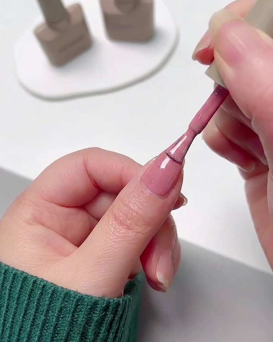 Une main appliquant une couche lisse de vernis gel rose sur un ongle à laide du pinceau du flacon de vernis.