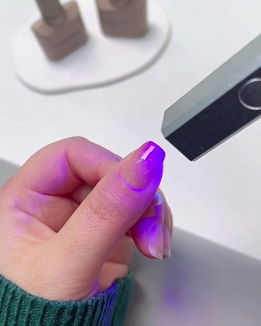 Un doigt positionné sous une petite lampe à ongles UV/LED portable émettant une lumière violette brillante pour catalyser le vernis gel.