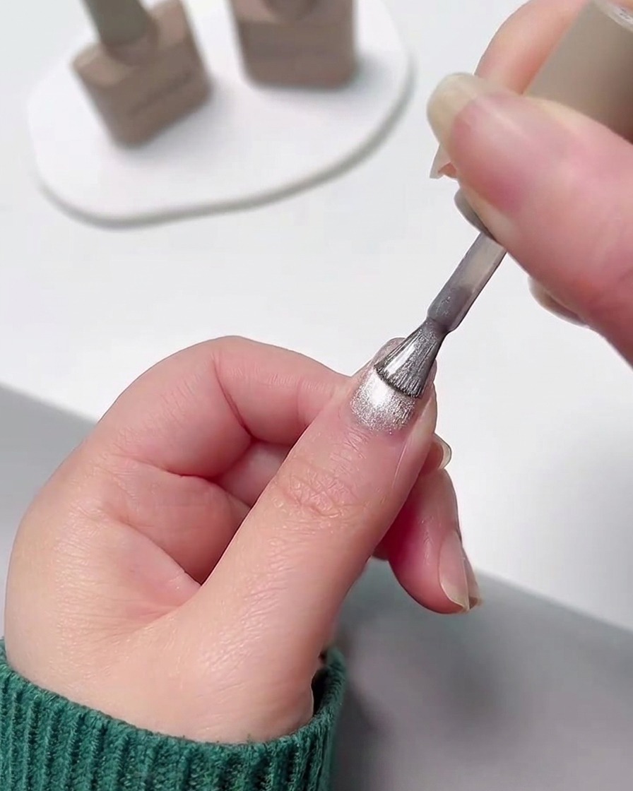 Une main appliquant une couche de vernis gel cat eye argenté scintillant sur un ongle au dégradé rose catalysé.