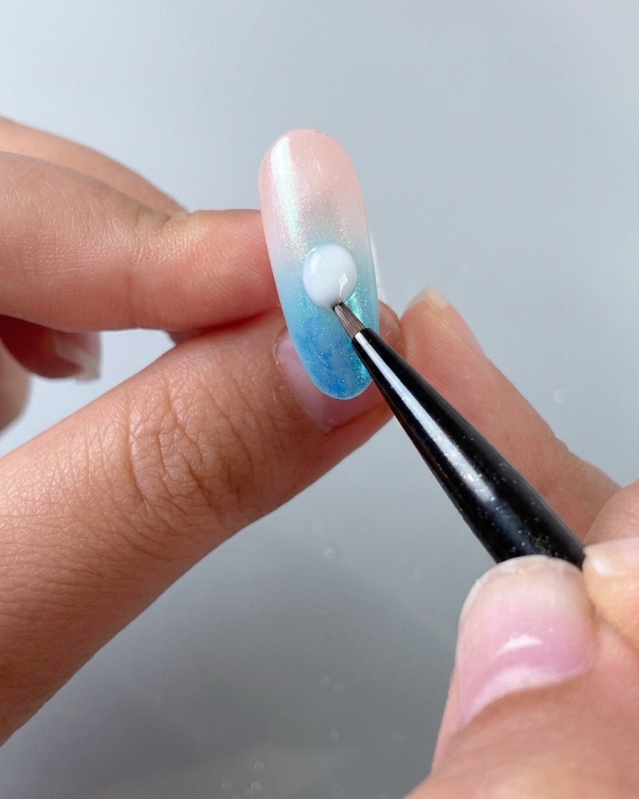 Un pinceau fin pour nail art appliquant une goutte de gel autonivelant blanc laiteux en diagonale sur un ongle en dégradé.