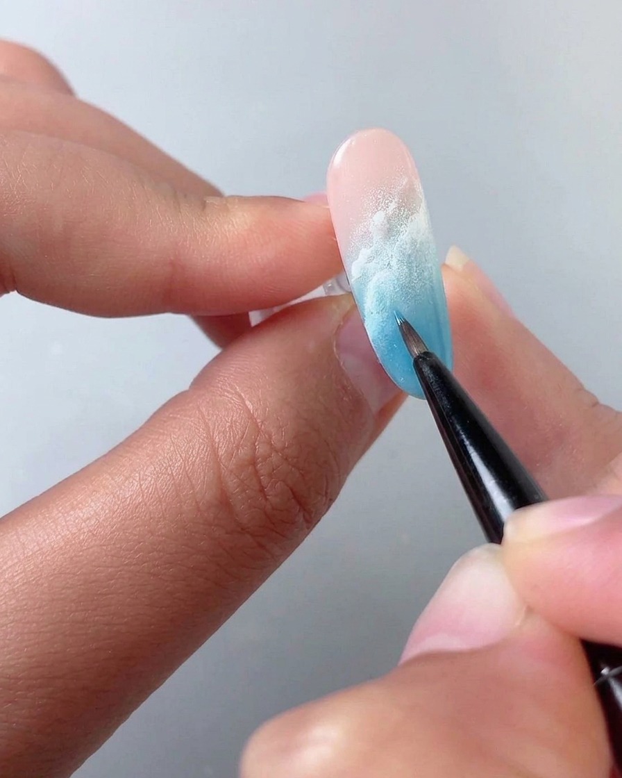 Un pinceau fin estompant du liquide blooming blanc sur un ongle en dégradé bleu et rose.