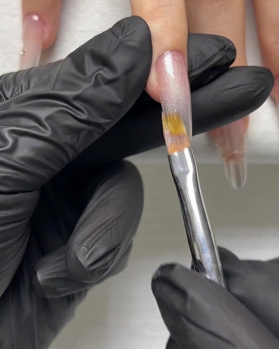 Main gantée utilisant un pinceau fin pour lisser une couche de gel scintillant argenté sur un ongle allongé.