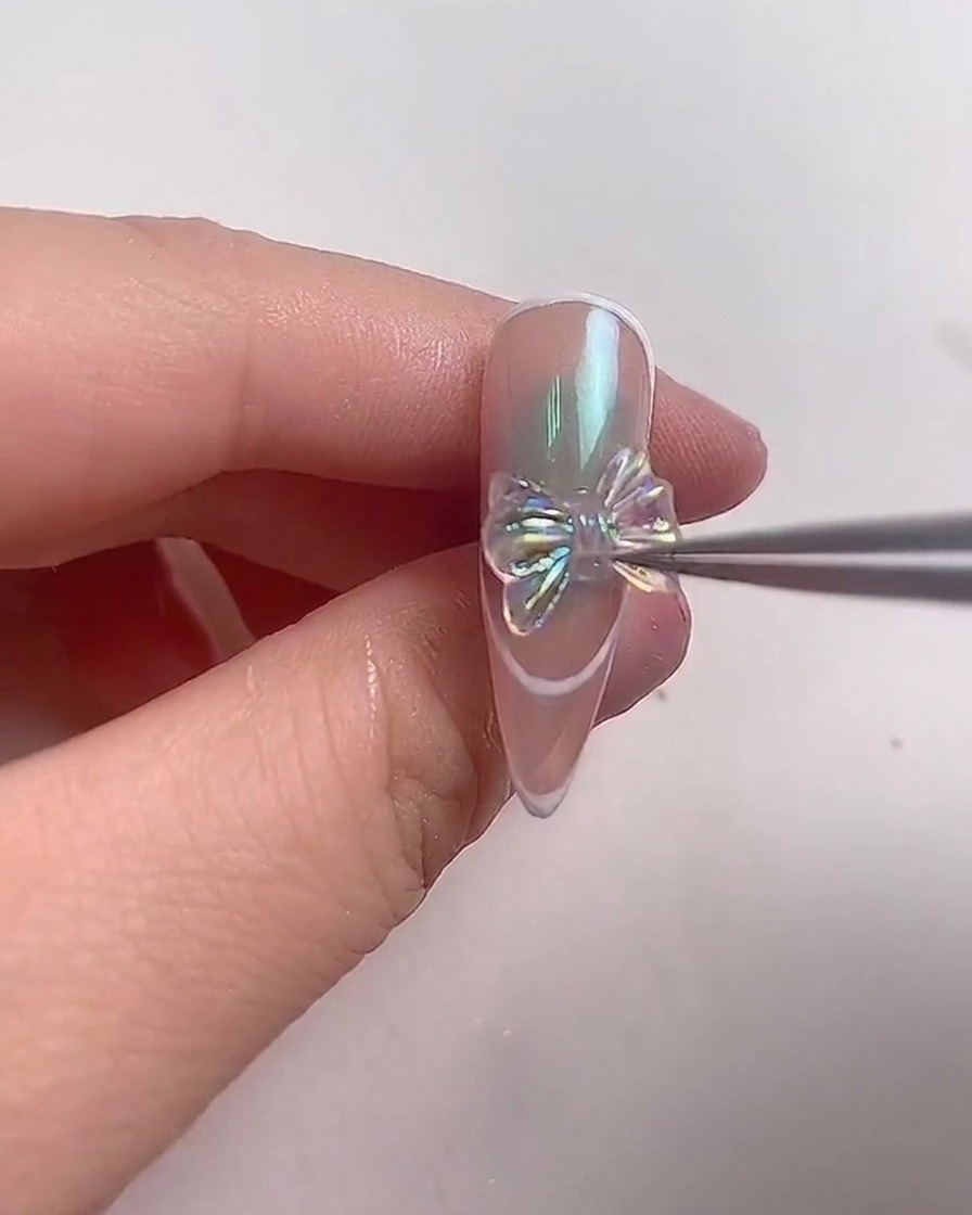 Une paire de pinces métalliques positionnant délicatement un nœud en cristal irisé au centre dun ongle en forme damande.