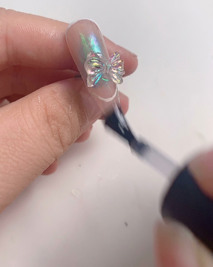 Un pinceau appliquant une dernière couche de top coat transparent autour dun bijou nœud en cristal sur un ongle terminé.