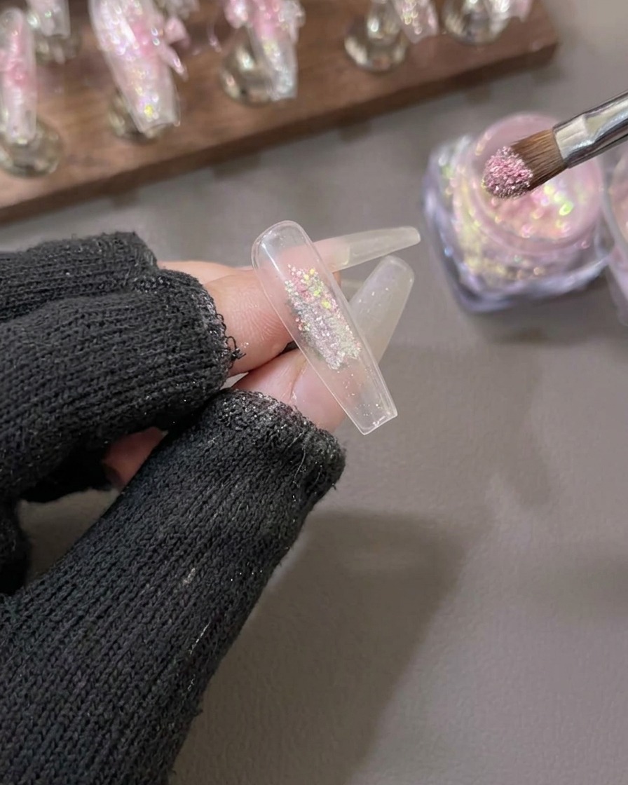 Un pinceau appliquant de la poudre de flocon aurora rose et dorée scintillante au centre dune capsule dongle artificielle transparente.