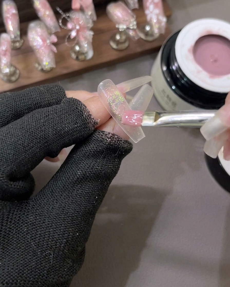 Un pinceau plat à gel étalant un vernis gel rose lait semi-transparent sur un ongle comportant des flocons pailletés au centre.