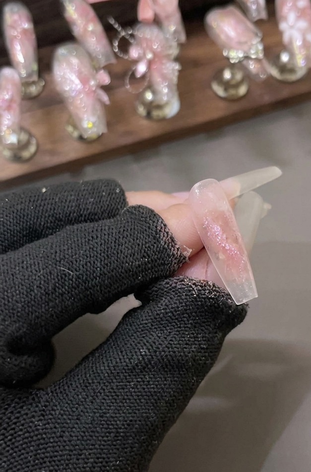 Une main gantée tenant un ongle transparent recouvert dune couche scintillante de gel à paillettes diamant brisé rose.
