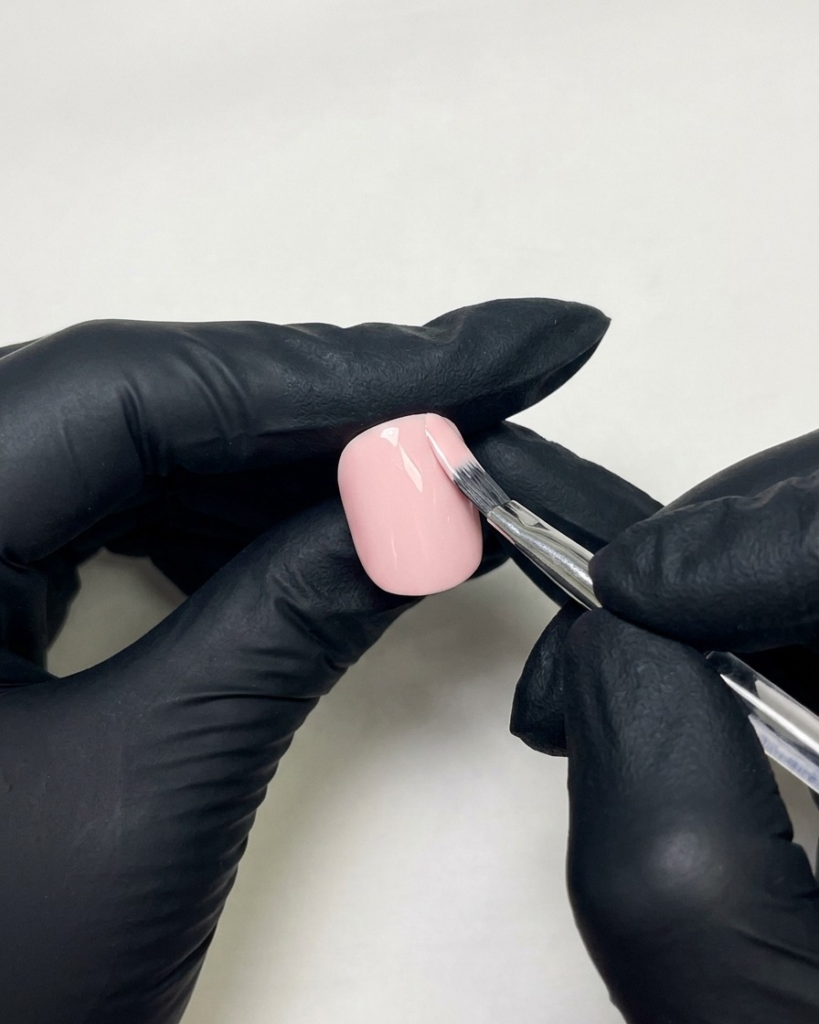 Mains gantées utilisant un pinceau plat pour appliquer du vernis gel rose peau sur un ongle préparé.
