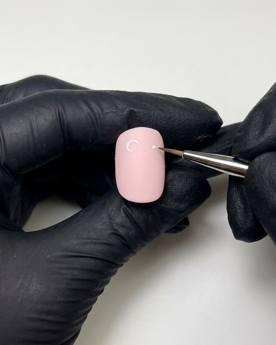Mains gantées utilisant un pinceau liner fin avec du builder gel pour dessiner une boucle et un point blanc-rose sur un ongle vernis.