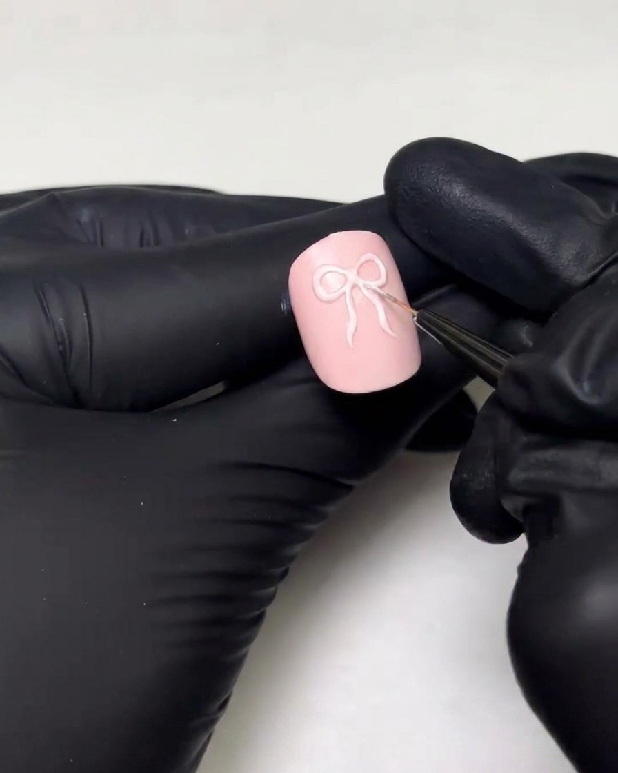 Une main gantée utilisant un pinceau de détail fin pour dessiner les boucles et les queues de ruban dun motif de nœud 3D sur un ongle rose.