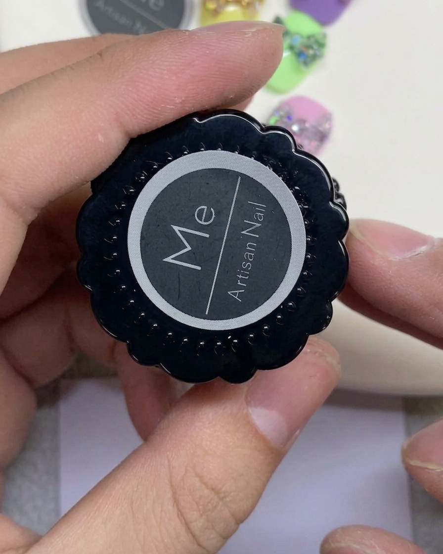 Une main tenant un pot noir étiqueté Artisan Nail avec un logo circulaire blanc.