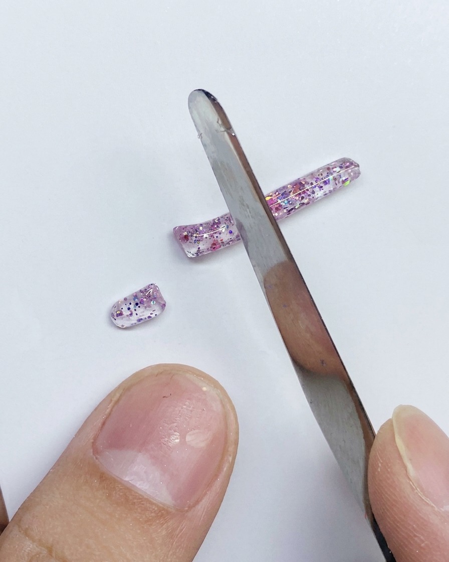 Une spatule en métal coupant une longue bande de gel PVC rose pailleté en segments plus petits sur une surface blanche.
