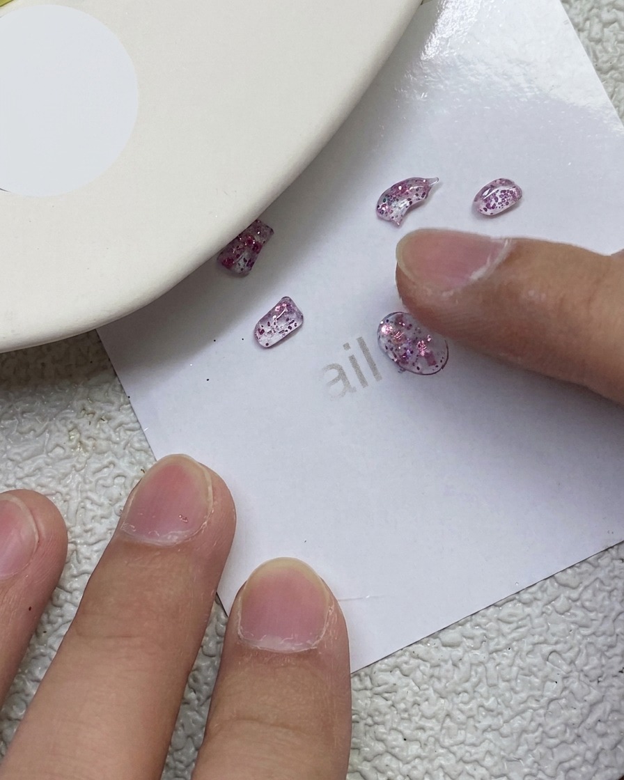 Un doigt pressant et sculptant de petits morceaux de gel PVC rose pailleté en forme de nœuds sur une feuille de papier blanc.