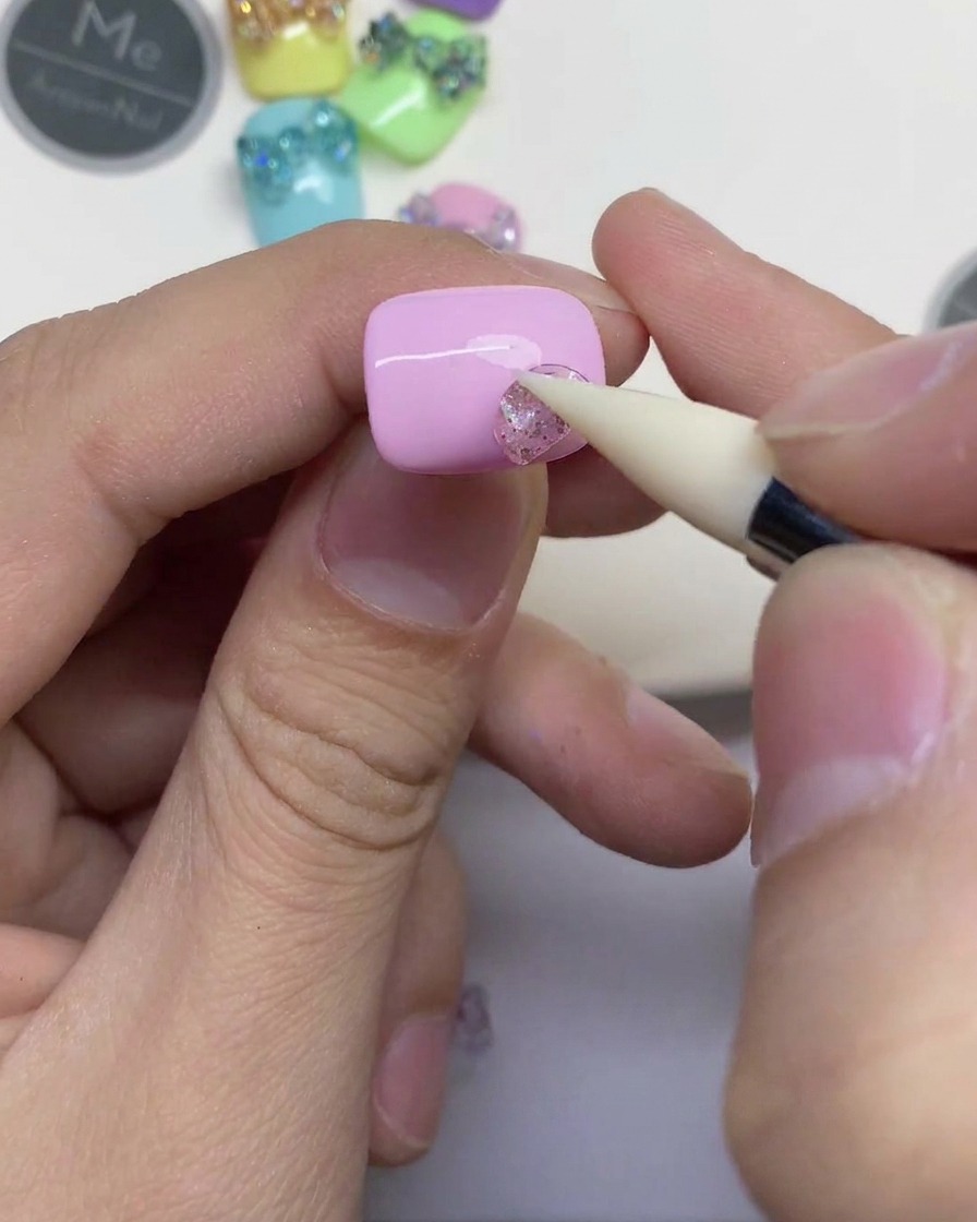 Gros plan dune main utilisant un outil en silicone blanc pour presser un petit morceau de gel pailleté transparent sur un ongle rose clair.