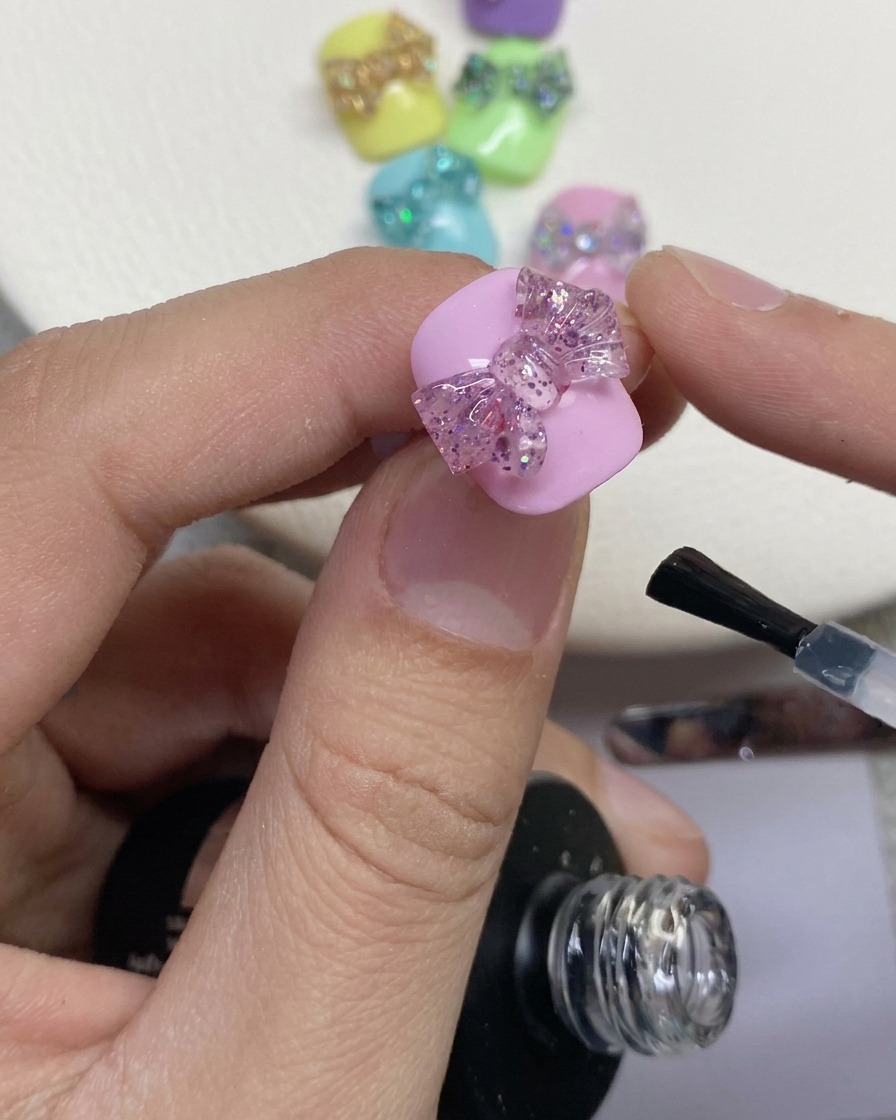 Gros plan dun pinceau à ongles appliquant une couche de finition transparente autour dun nœud en paillettes 3D sur un ongle peint en rose.