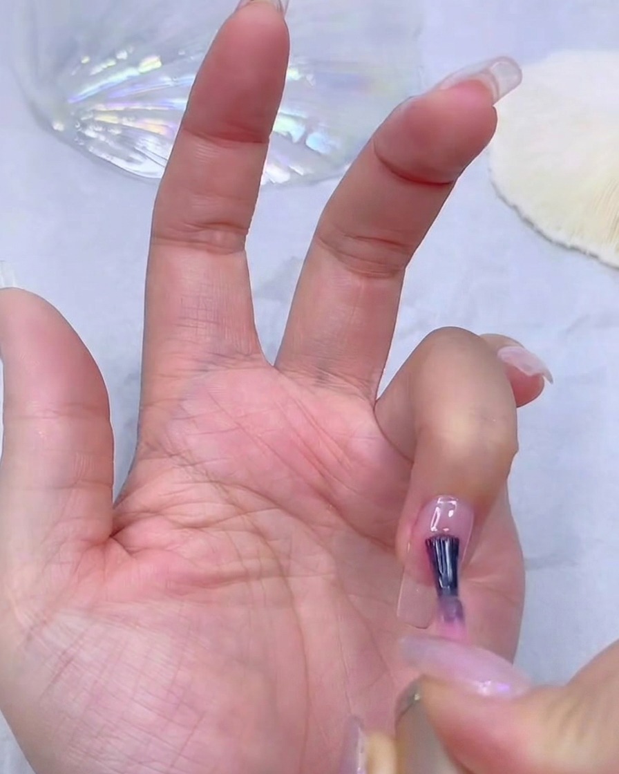 La main dune prothésiste ongulaire appliquant une fine couche de base gel rose sur une extension dongle transparente.