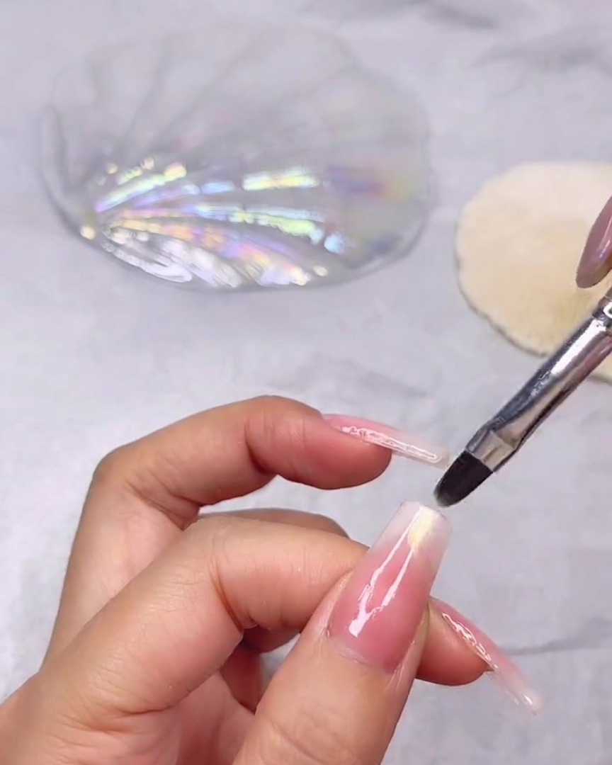 Un pinceau fin appliquant un gel transparent nacré effet coquillage sur la pointe dun ongle long orné dun dégradé rose et blanc.