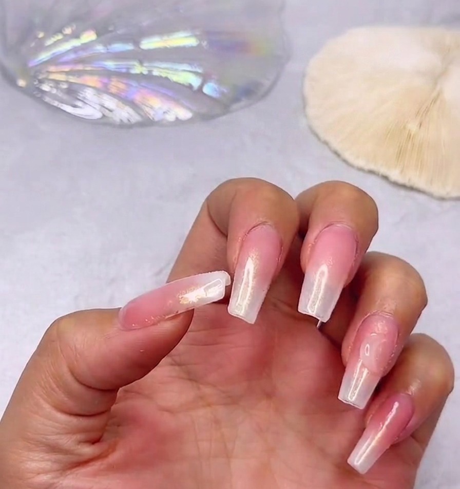 Gros plan dune main arborant des ongles longs brillants avec un dégradé rose et blanc laiteux, ornés dun grand strass cœur transparent placé près de la cuticule du pouce.