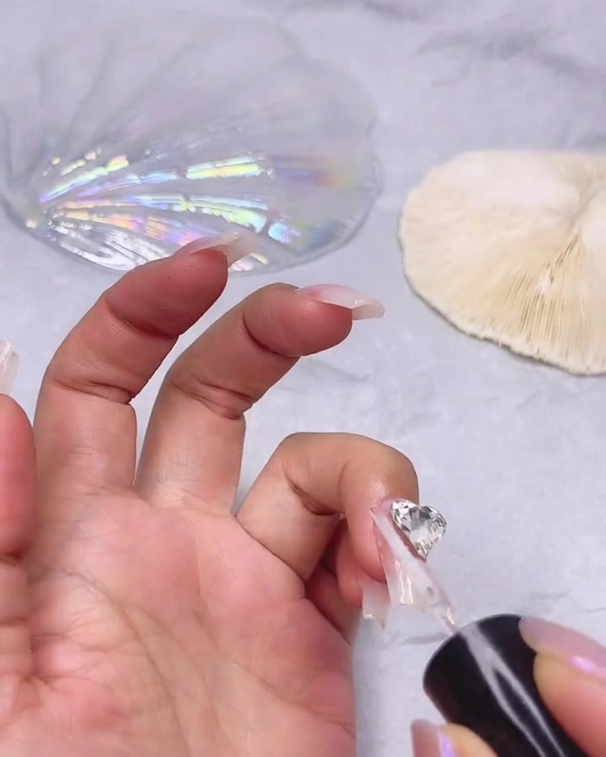 Un pinceau fin appliquant un gel de scellement transparent autour des bords dun grand strass cœur fixé sur un ongle.