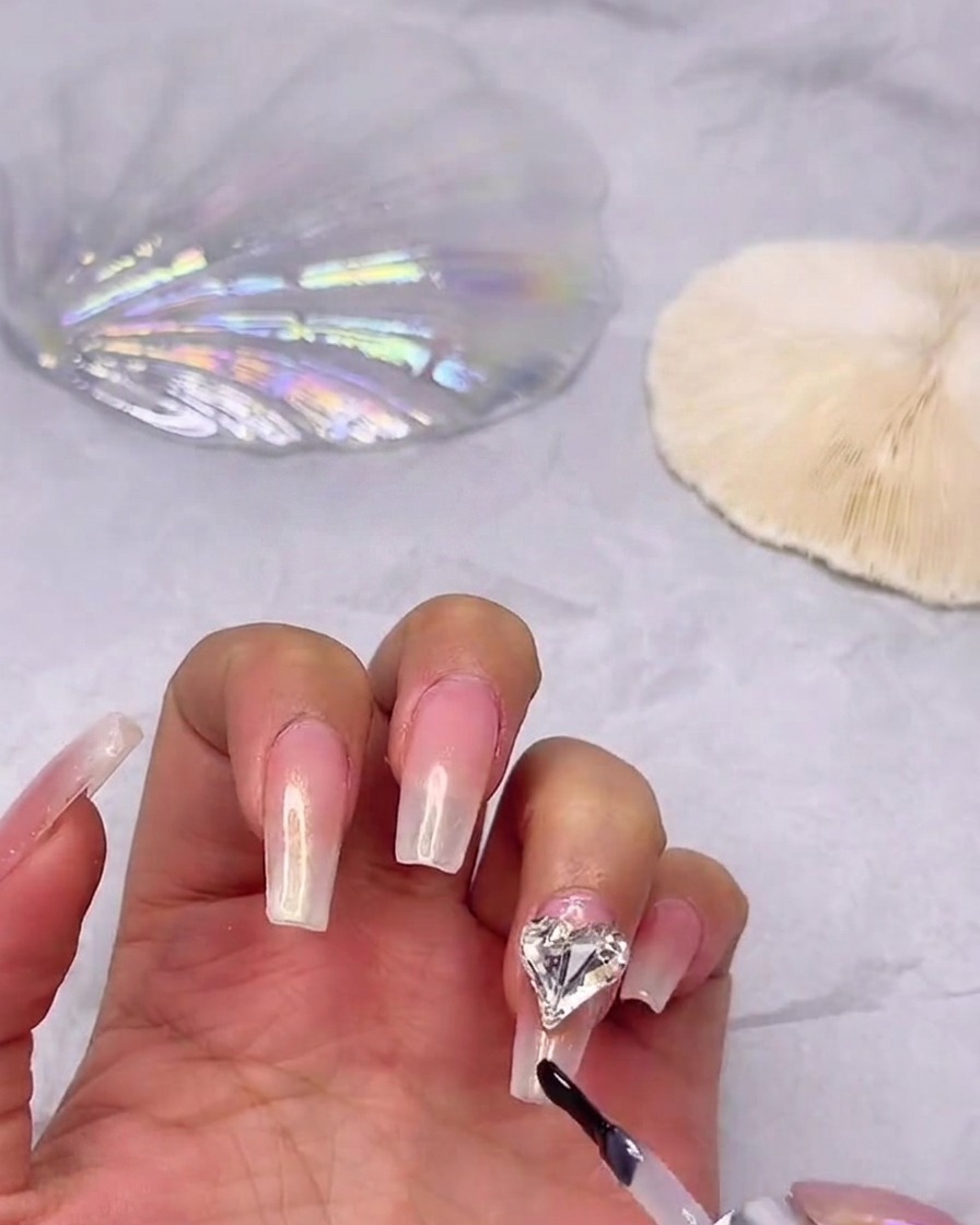 Un pinceau de vernis gel appliquant un top coat brillant sur un ongle dégradé, en contournant soigneusement un strass cœur fixé.