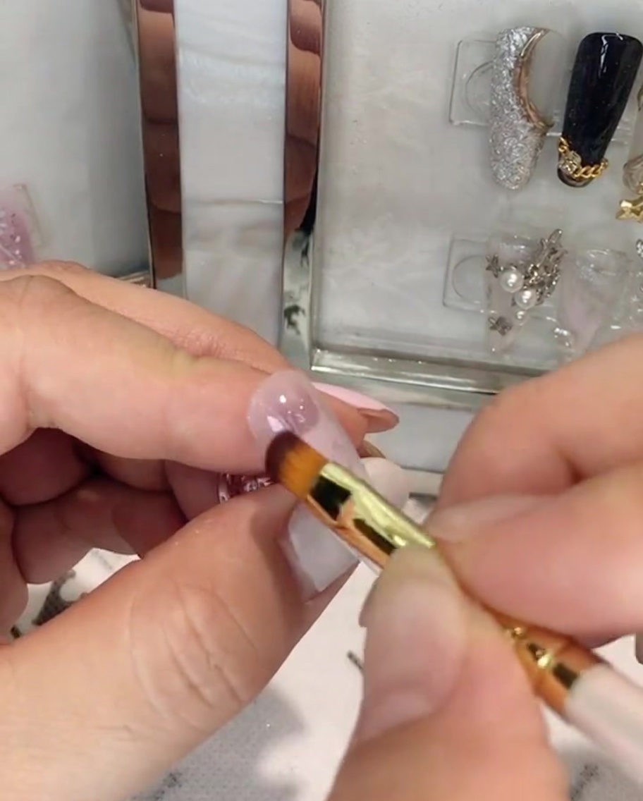 Une main tenant un pinceau à ongle appliquant du gel rose sur la moitié supérieure dun ongle en amande.