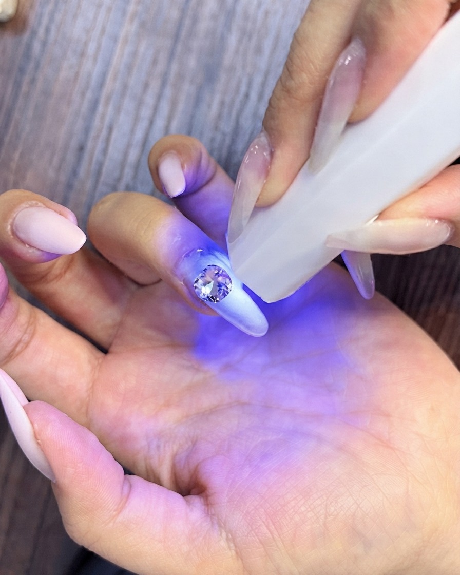 Gros plan dune main ramassant de petits strass en cristal pour les agencer autour dune pierre centrale plus grande sur un ongle manucuré.