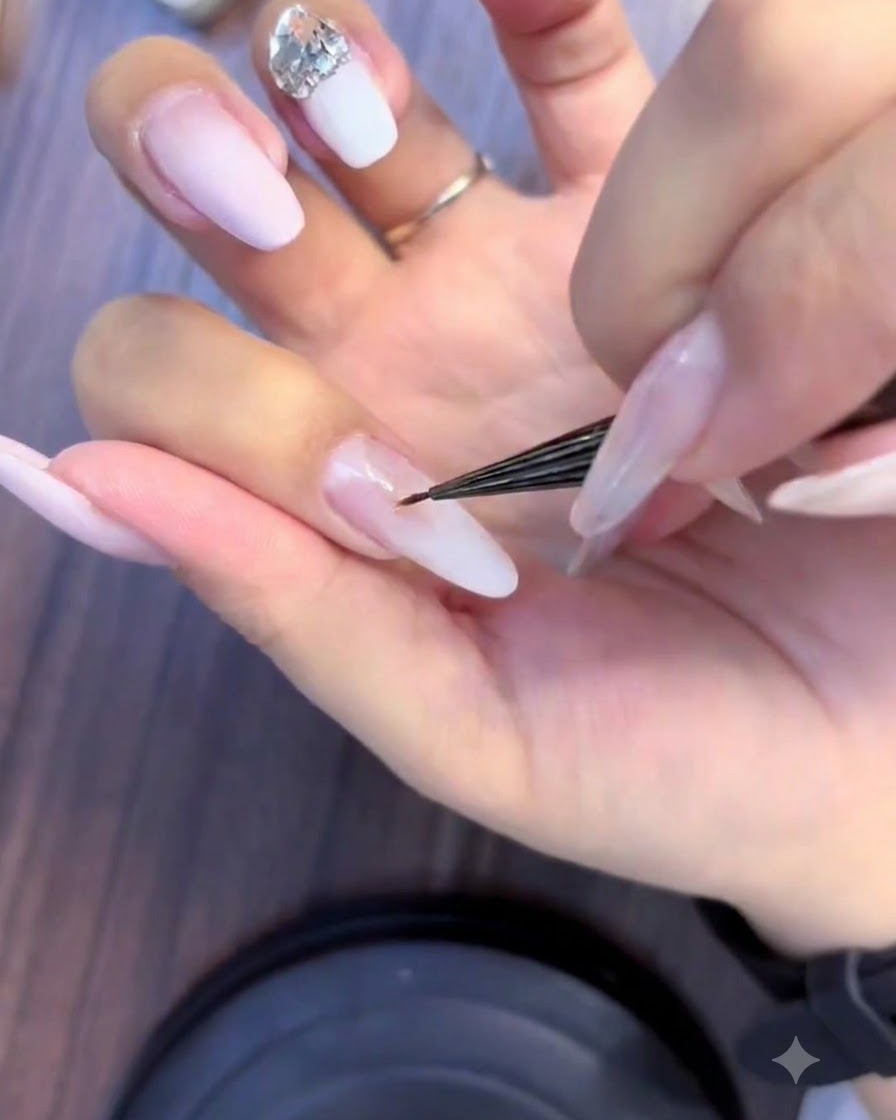 Gros plan dune prothésiste ongulaire utilisant un pinceau fin pour appliquer de petits strass dans une grappe de cristaux sur un ongle ombré rose et blanc.