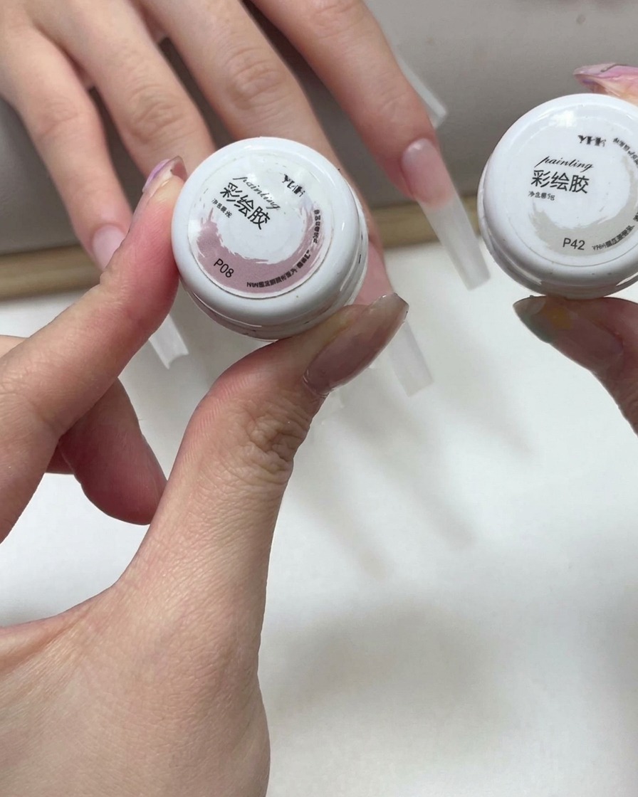 Mains tenant deux petits pots de gel de couleur rose et blanc avec des ongles allongés visibles en arrière-plan.
