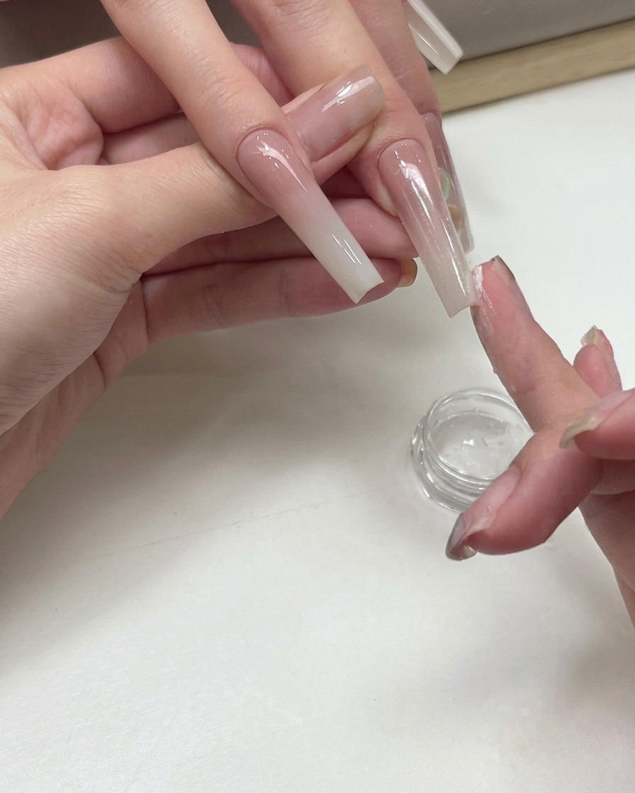 Une prothésiste ongulaire utilisant le bout de son doigt pour tapoter de la poudre de pigment scintillante pêche blanche sur la surface dun ongle étendu en dégradé.