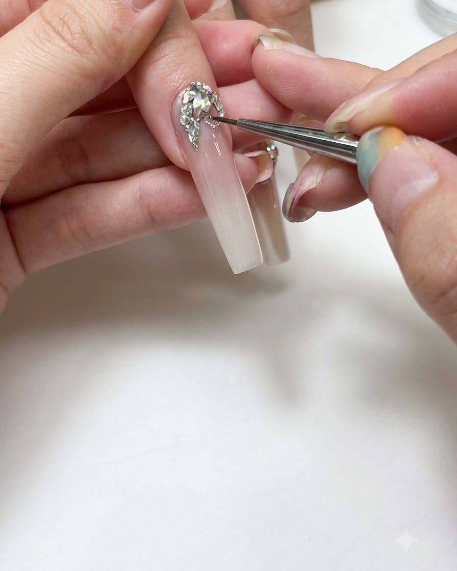 Gros plan dune prothésiste ongulaire utilisant un pinceau de détail fin pour peindre des lignes en gel argenté autour de cristaux transparents sur un long ongle ombré et étendu.