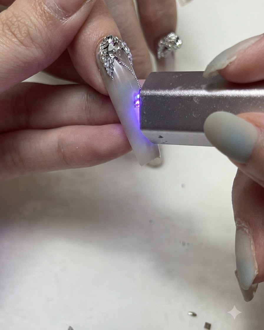 Une lampe torche UV/LED portable catalysant de fines lignes de gel argenté et des détails de strass sur un ongle décoré.