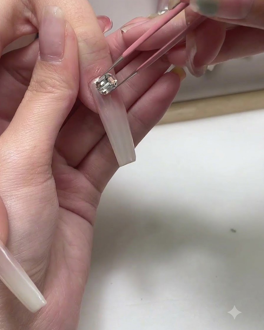 Une paire de pinces plaçant un petit charm métallique sur un long ongle en gel rose nude.