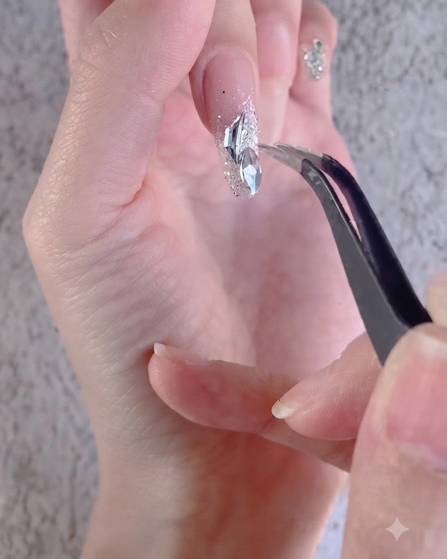 Une main utilisant une pince pour agencer des strass blancs irréguliers sur un ongle décoré.