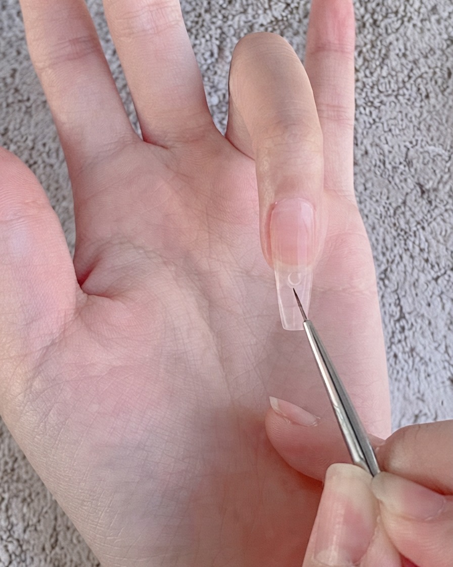 Un pinceau liner fin de nail art déposant une petite perle transparente de gel à strass sur un ongle recouvert de gel nude.