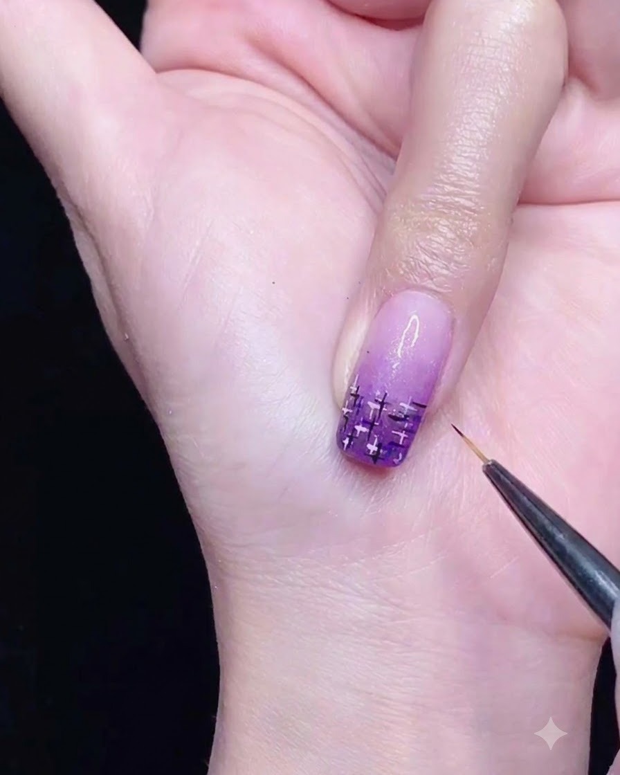 Un pinceau liner fin dessinant des lignes noires et argentées qui sentrecroisent sur un ongle dégradé violet.