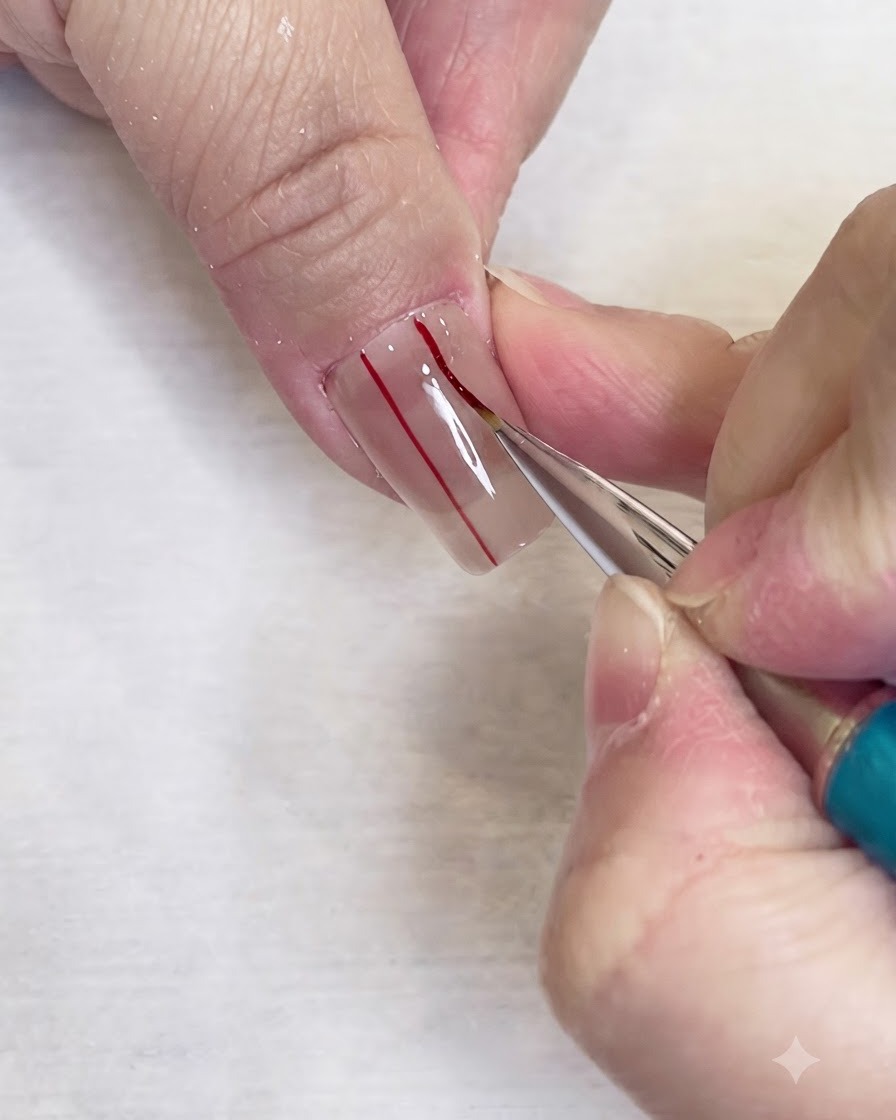 Un pinceau liner fin dessinant une seconde ligne verticale rouge parallèle sur un ongle transparent.