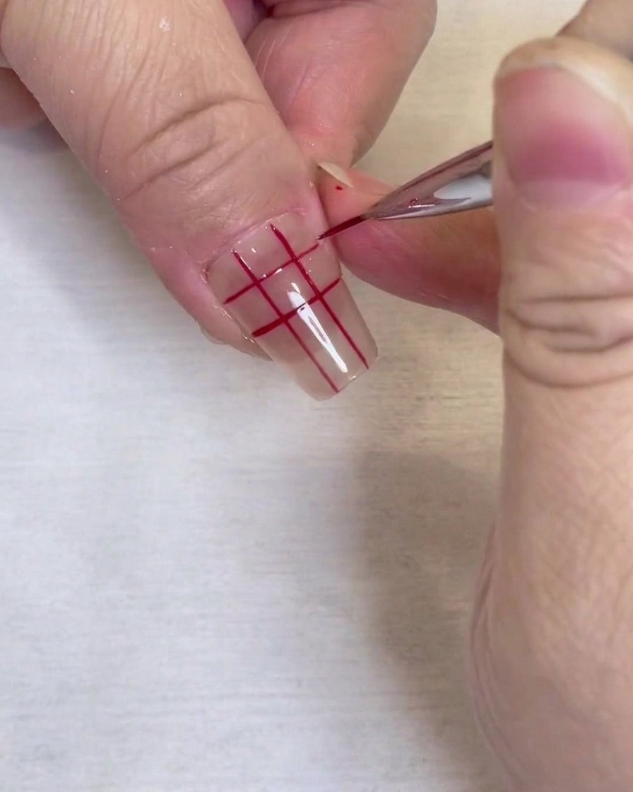 Gros plan dun motif de grille rouge terminé sur un ongle transparent, dessiné avec un pinceau liner fin.