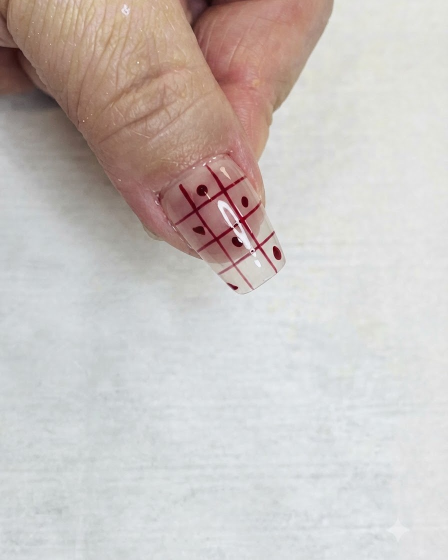 Un pinceau liner fin remplissant les carrés alternés dun motif de grille rouge sur un ongle transparent avec du vernis rouge.