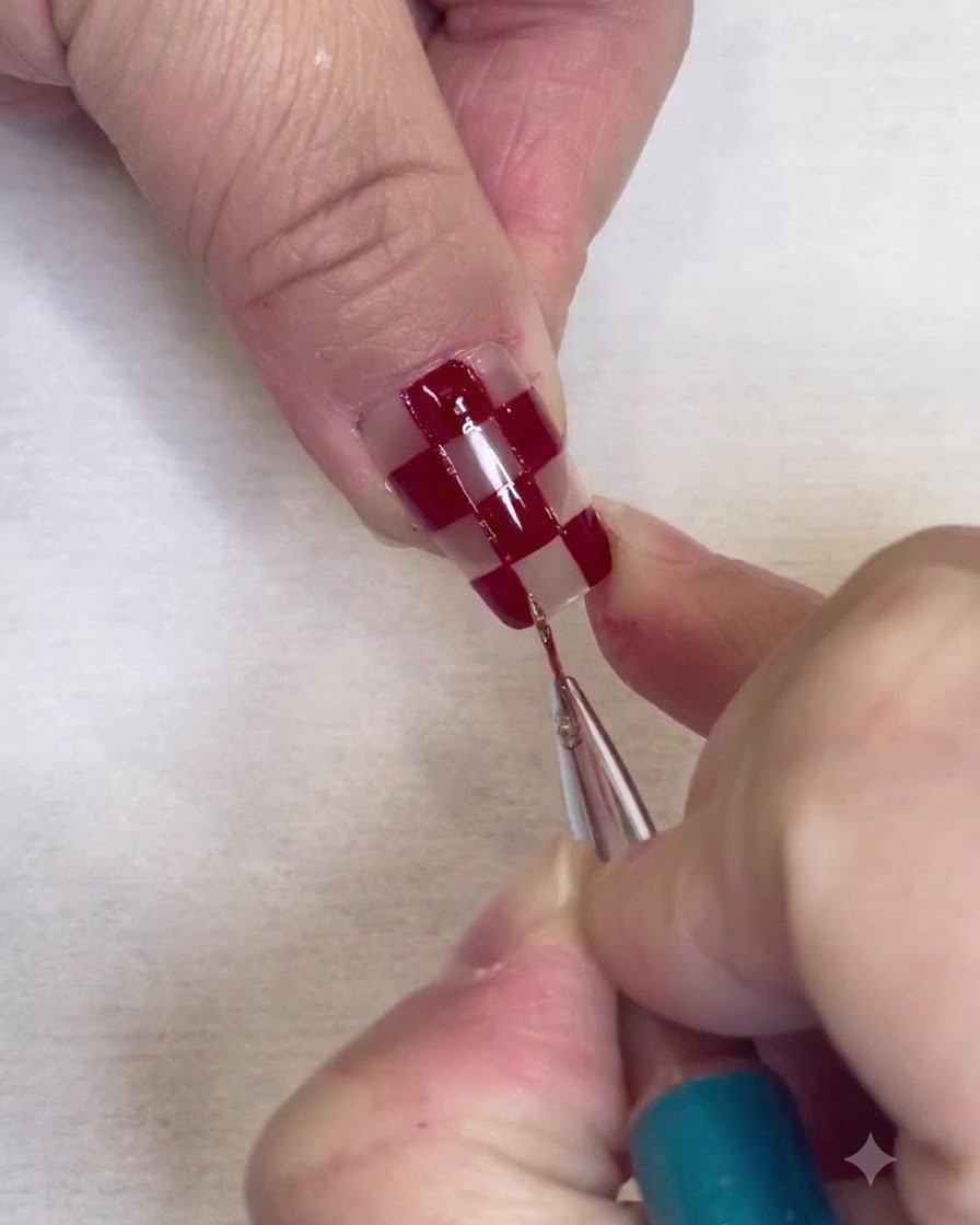 Un pinceau de détail fin peignant de fines bordures de paillettes argentées le long des bords des carrés rouges sur un ongle transparent.