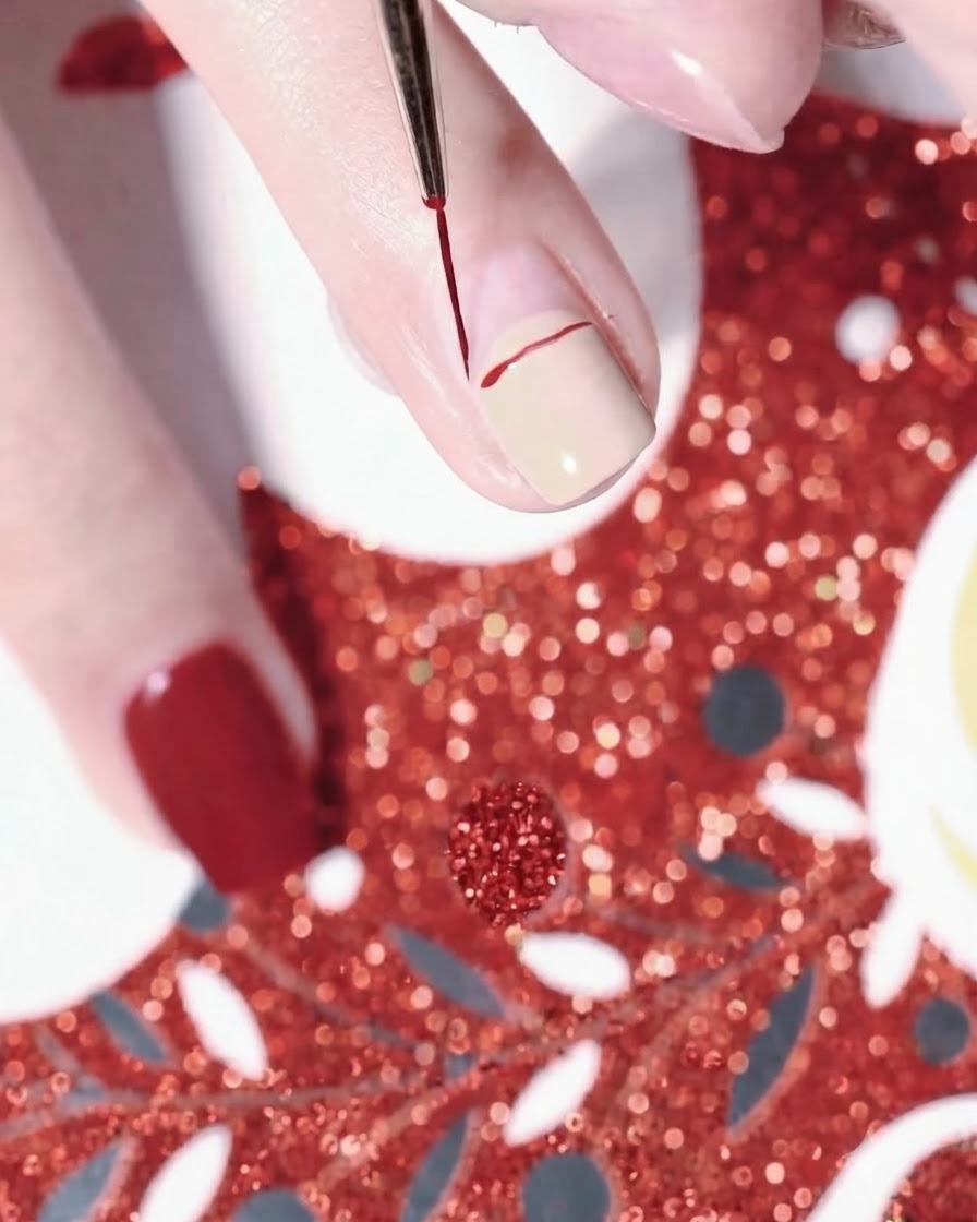 Gros plan dune main utilisant un pinceau liner fin pour dessiner la ligne courbe rouge dun bonnet de Père Noël sur un ongle peint en nude.