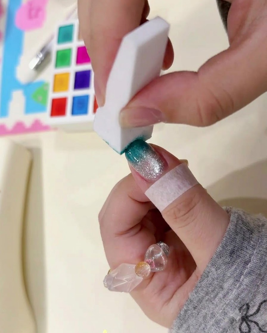 Une éponge blanche tapotant doucement le bout de longle pour créer un dégradé bleu canard foncé.