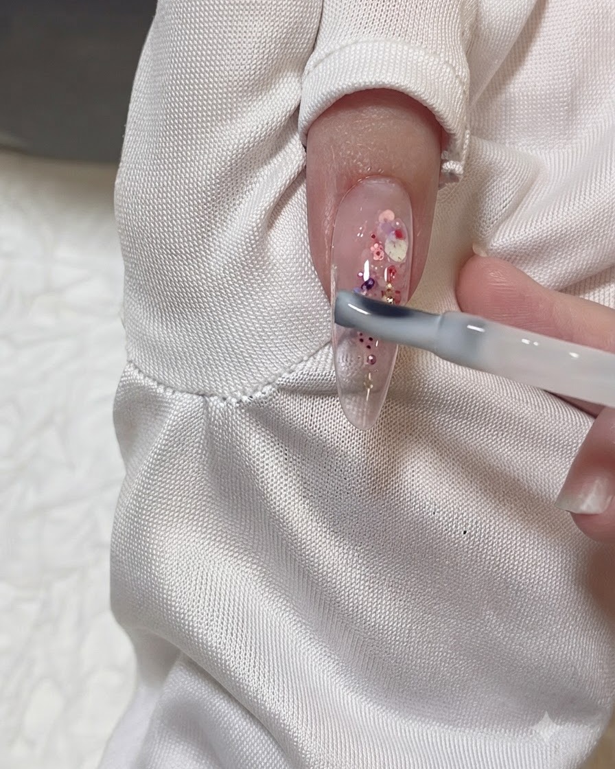 Un pinceau à gel appliquant une fine couche de gel lacté transparent sur un ongle amande transparent décoré de paillettes roses et violettes.