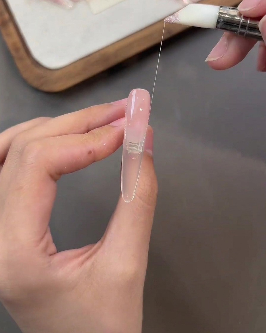 Un pinceau tirant un fin fil de gel rose pailleté sur la surface dun ongle de couleur nude.