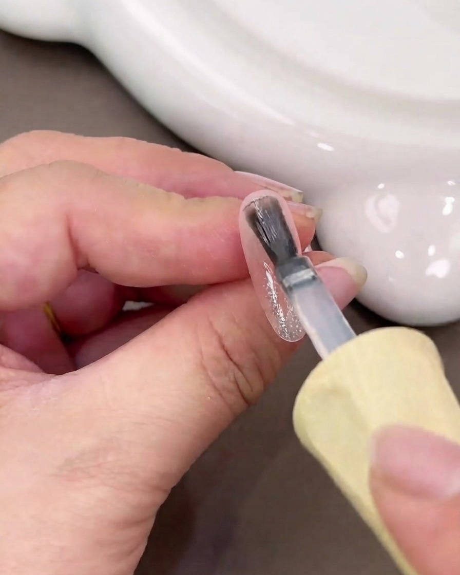 Un pinceau à ongles appliquant une couche humide de gel de base transparent sur une surface dongle brillante.