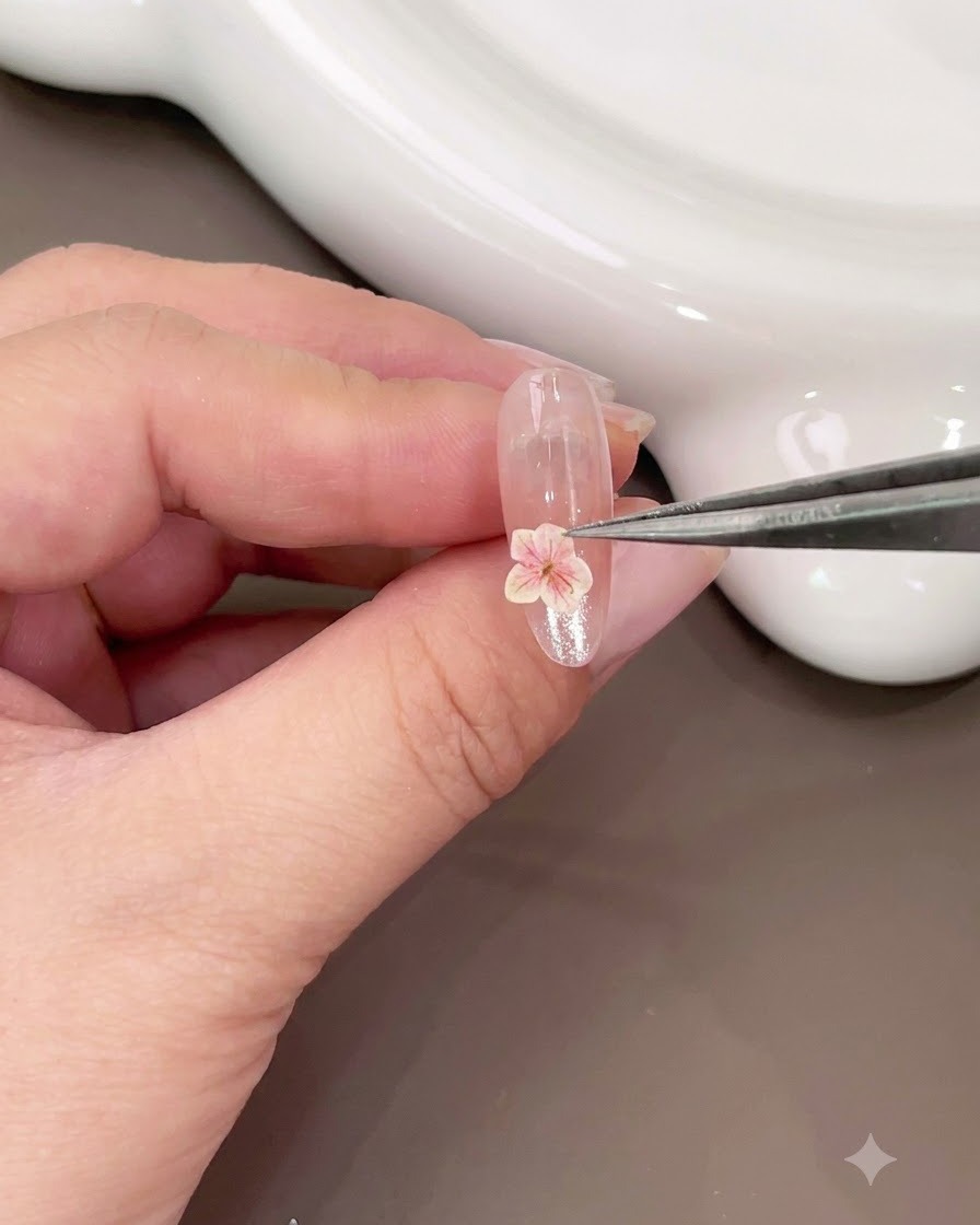 Une pince à épiler posant une petite fleur séchée rose sur la partie inférieure dun ongle brillant.